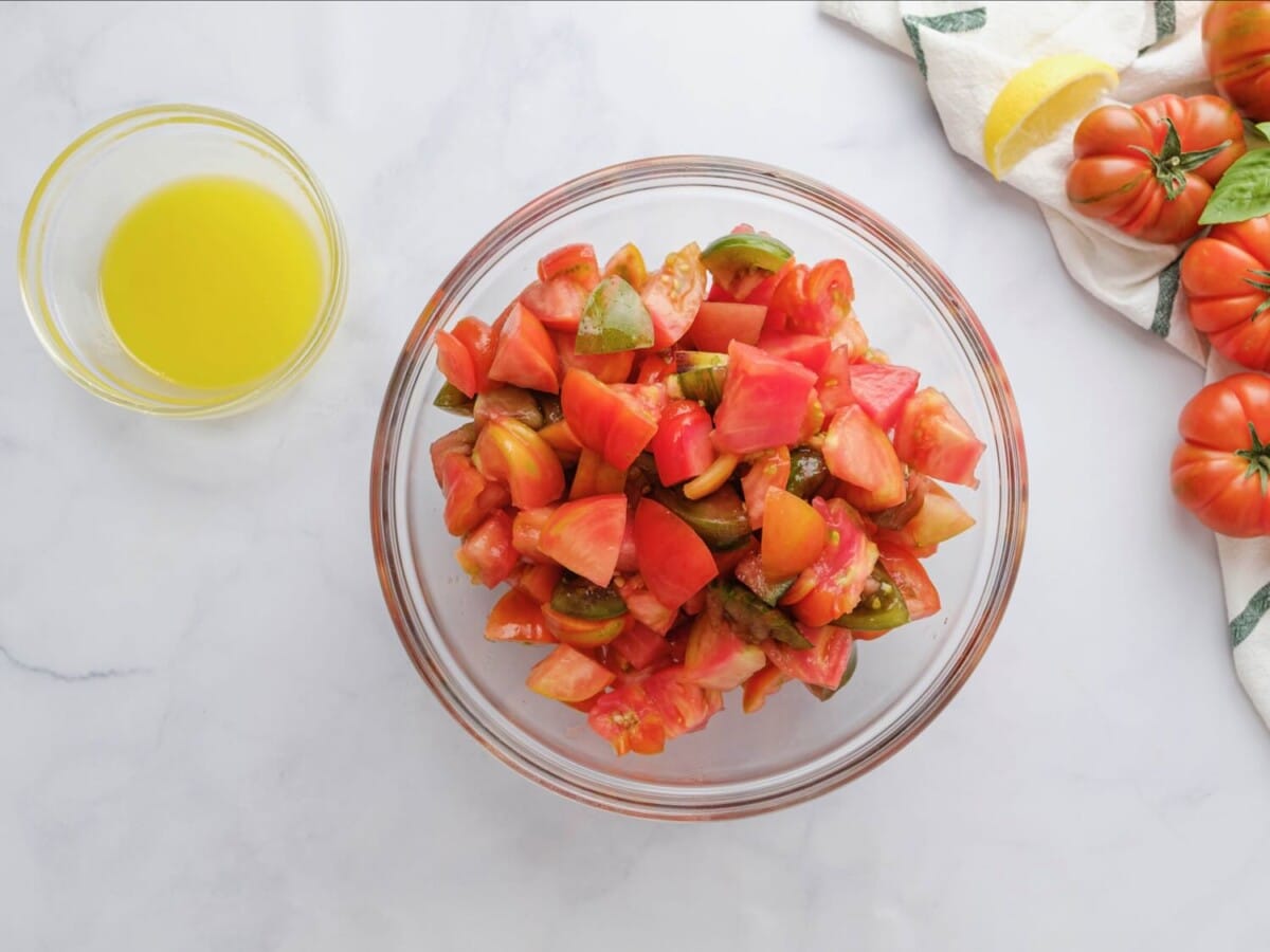 Glass bowl of chopped heirloom tomatoes with a small bowl of lemony olive oil dressing on the side, on a white marble background. Colorful heirloom tomatoes off to the right side, with a cloth napkin and lemon slices.