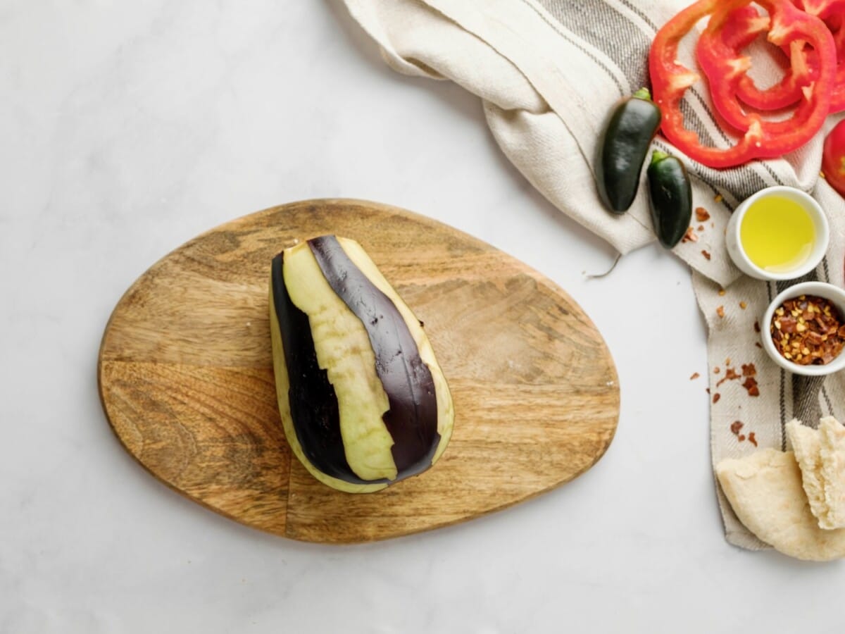 Medium eggplant with stem removed, peeled in strips so that part of the peel remains, while part of the peel is missing in long strips lengthwise down the eggplant. Linen napkin, small green peppers, red bell pepper slices, and chili flakes alongside the pot on the counter.