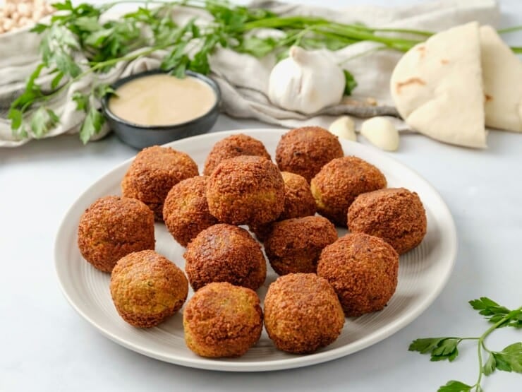 A white plate filled with falafel balls, tahini sauce, fresh herbs,a head of garlic, and pita bread sit in the background.