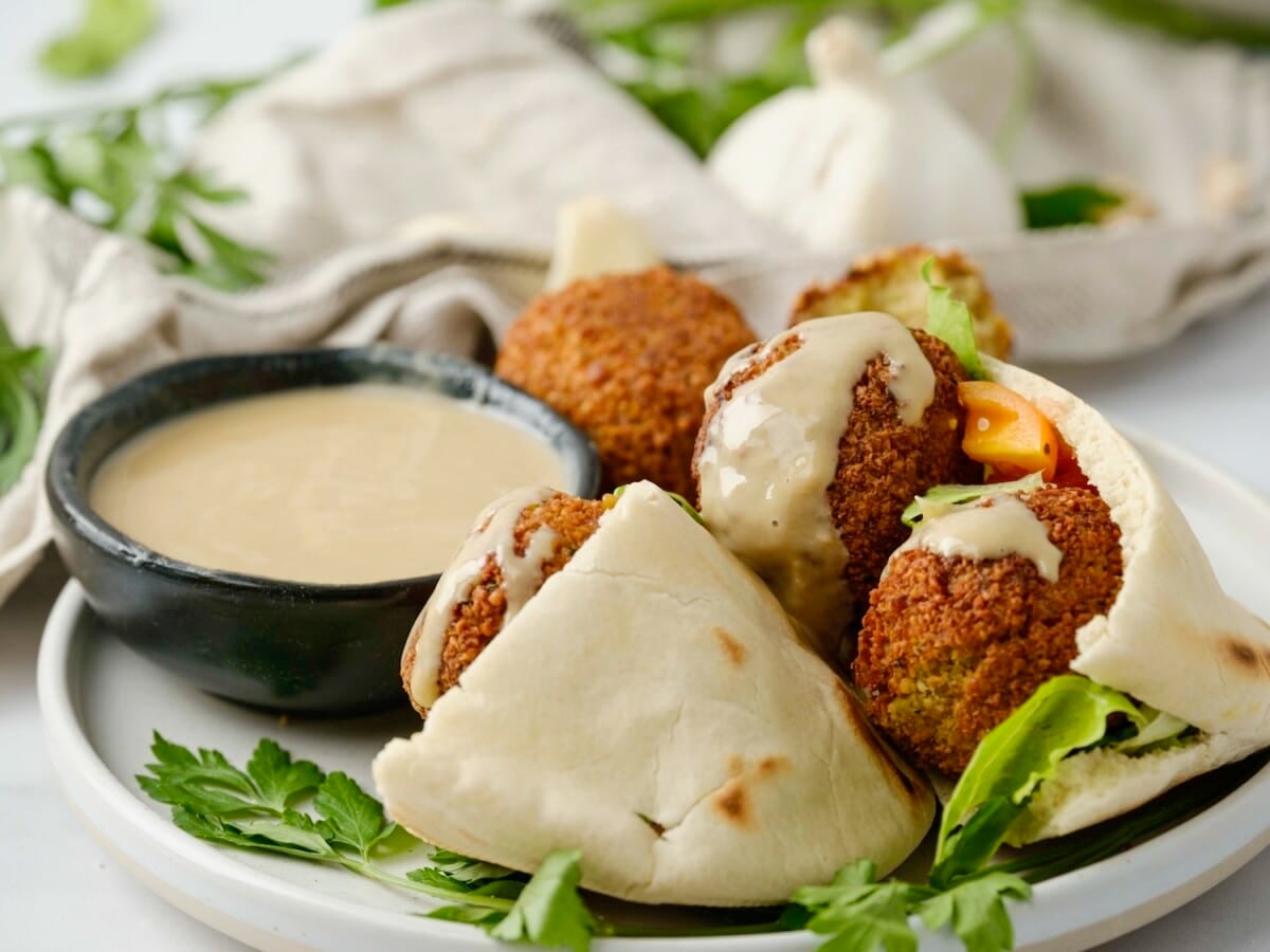 Horizontal close up image of a plate containing a pita pocket filled with falafel next to a small black dish of tahini sauce.