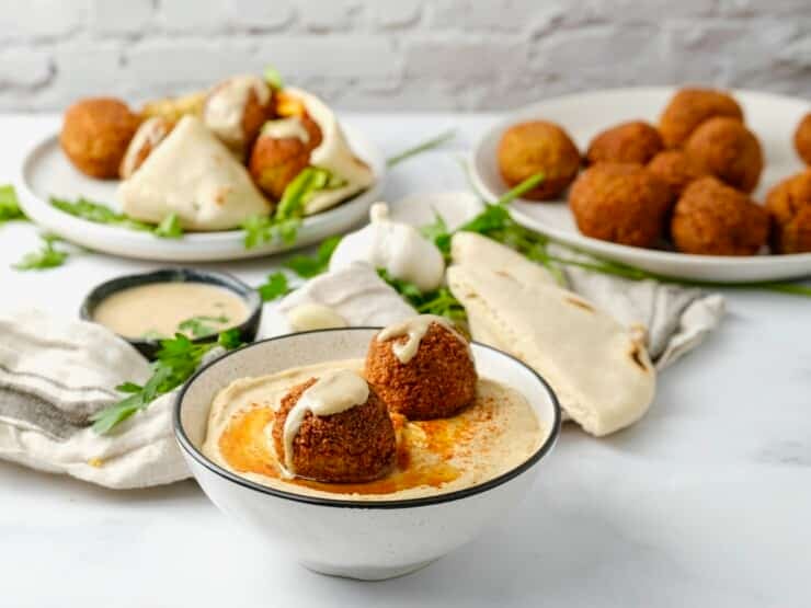 A bowl of hummus topped with two falafel balls. A plate of falafel balls sits in the background. 