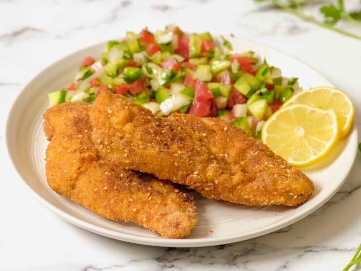 A plate of chicken schnitzel, chopped salad in background with lemon, on marble counter.