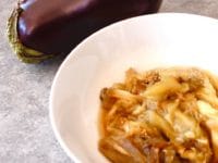 Roasted eggplant pulp in bowl with whole eggplant in background on grey background