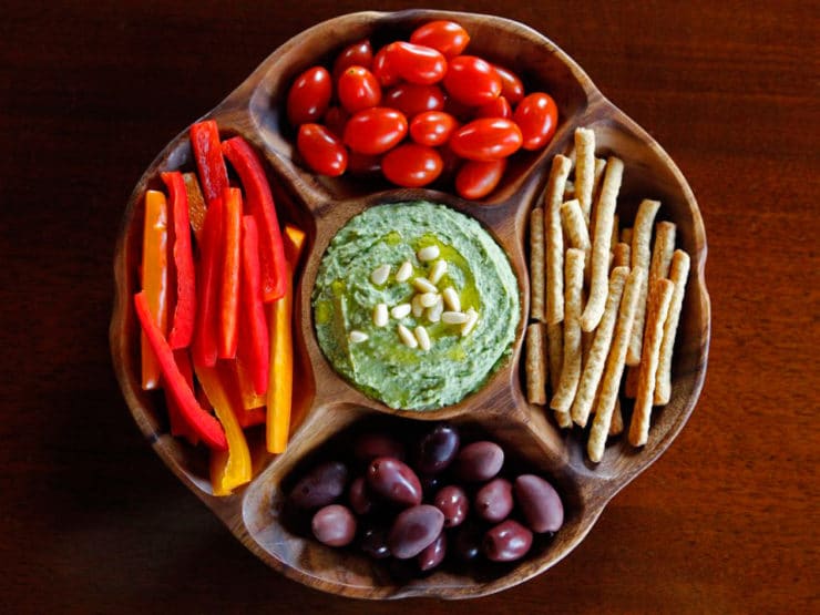 White bean basil pesto hummus on a platter with grape tomatoes, crackers, olives, and sliced bell peppers. 