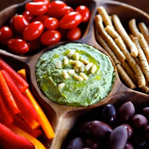 White bean basil pesto hummus on wooden plate, surrounded by pine nuts, bell pepper, grape tomatoes, and breadsticks.