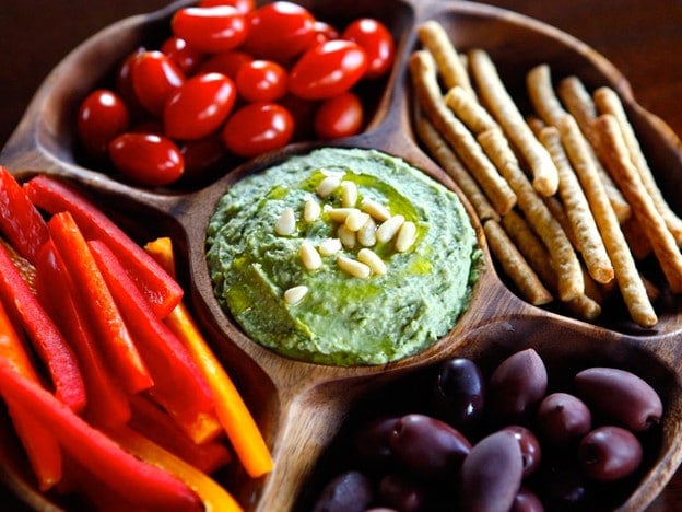 White bean basil pesto hummus on wooden plate, surrounded by pine nuts, bell pepper, grape tomatoes, and breadsticks.
