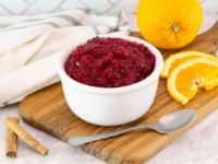 Boozy Orange Cranberry Sauce Pinterest Pin - a bowl of cranberry sauce on a wooden board, surrounded by sliced and whole oranges, cinnamon sticks, a spoon and a towel is draped beside it.