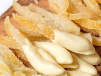 Horizontal shot - wide shot of candied lemon peels on a small wood cutting board on marble counter. Large peels, small peels, peels dipped in white chocolate. Lemon half and white linen towel in background.