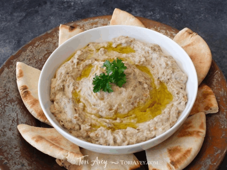 A bowl of baba ganoush topped with olive oil and herbs surrounded by pita bread. 