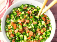 A white a bowl filled with mixed cucumbers, bell peppers, onions, and diced tomatoes and wooden spoons for mixing
