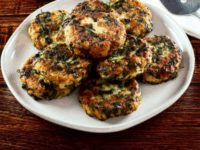 Keftes de Espinaca Spinach Keftes Pinterest Pin - A plate of savory Keftes de Espinaca - Spinach Keftes stacked on a wooden table, next to fresh spinach leaves and a jar of salt. A striped kitchen towel is also visible.