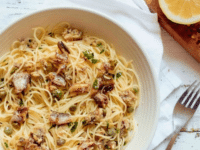 White bowl of sardine pasta capellini garnished with chili flakes and fresh parsley, resting on a white cloth napkin, wooden cutting board in background with lemon half and chili flakes, on white wood table.