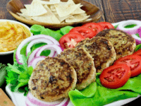 Close-up of four Mediterranean turkey burgers served with chopped tomatoes, onions, lettuce, chips, and a dipping sauce
