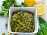 Close-up of Pistachio Mint Pesto in a small bowl surrounded with mint, basil leaves, pistachio, lemons, and garlic