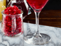 Pomegranate Martini Pinterest Pin - Martini glass with pomegranate martini on marble cutting board, glass carafe of pomegranate seeds, glass carafe of lime juice, martini shaker in background, on dark wooden surface.