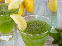 Two glasses of spiked Limonana served with a slice of lemon garnish surrounded with lemons, a tall fancy glass, and mint leaves in the background