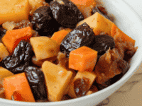 Horizontal shot - white bowl of tzimmes with sweet potatoes, prunes, carrots, and dried cranberries on a marble countertop, linen napkin with dried cranberries in background.