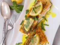 Two pieces of cooked salmon topped with lemon slices, fresh mint, and a buttery sauce on a white plate on a white background.